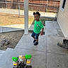 black_shoes, boot, child, concrete_porch, curious, daytime, denim_shorts, fence, grass, green_shirt, house, outdoor, sidewalk, standing, sunlight, toddler, toy_castle, tree, window, yard