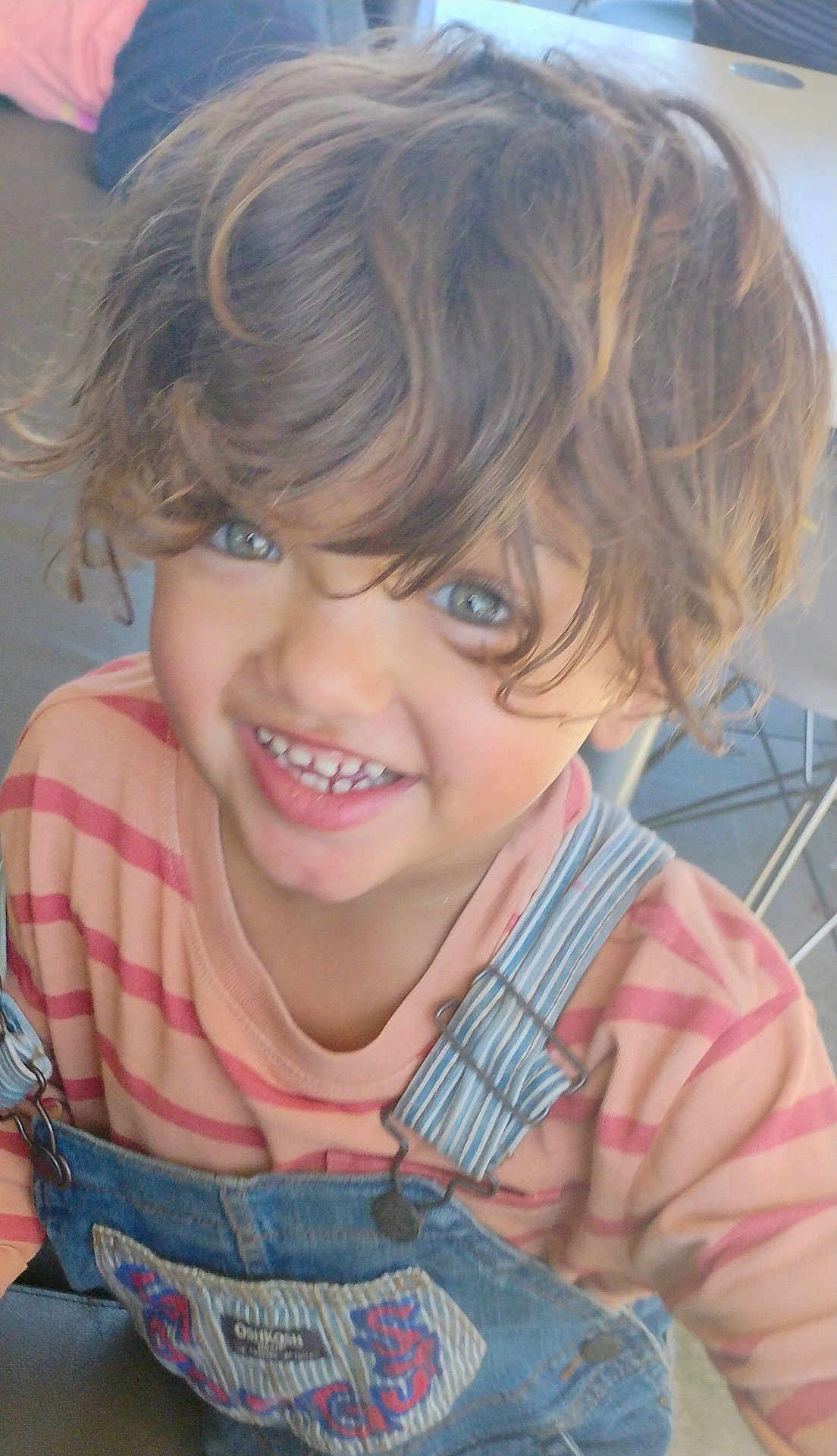 Harley is registered to the contest to win money with this photo: child, smiling, face, hair, denim_overalls, striped_shirt, indoor, table, chair, happy, cute, portrait, young, person, clothing, casual, toddler, eyes, expression, playful