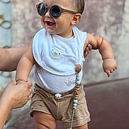Jude is registered to the contest to win money with this photo: baby, child, infant, sunglasses, smiling, laughing, portrait, standing, adult_hands, bib, onesie, shorts, pacifier, pacifier_clip, clothing, indoor, photography, happy, fashionable, closeup