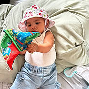 Adaline is registered to the contest to win money with this photo: baby, child, hat, sunhat, jeans, white_tank_top, fabric_book, bed, pillow, striped_bed_sheet, indoor, infant, cute, play, toy, colorful, relaxed, comfort, person, young