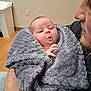 baby, blanket, person, face, indoor, adult, holding, expression, cozy, fur_texture, human_hand, tattoo, piercing, clothing, wood_floor, wall_socket, furniture, closeup, portrait, tenderness