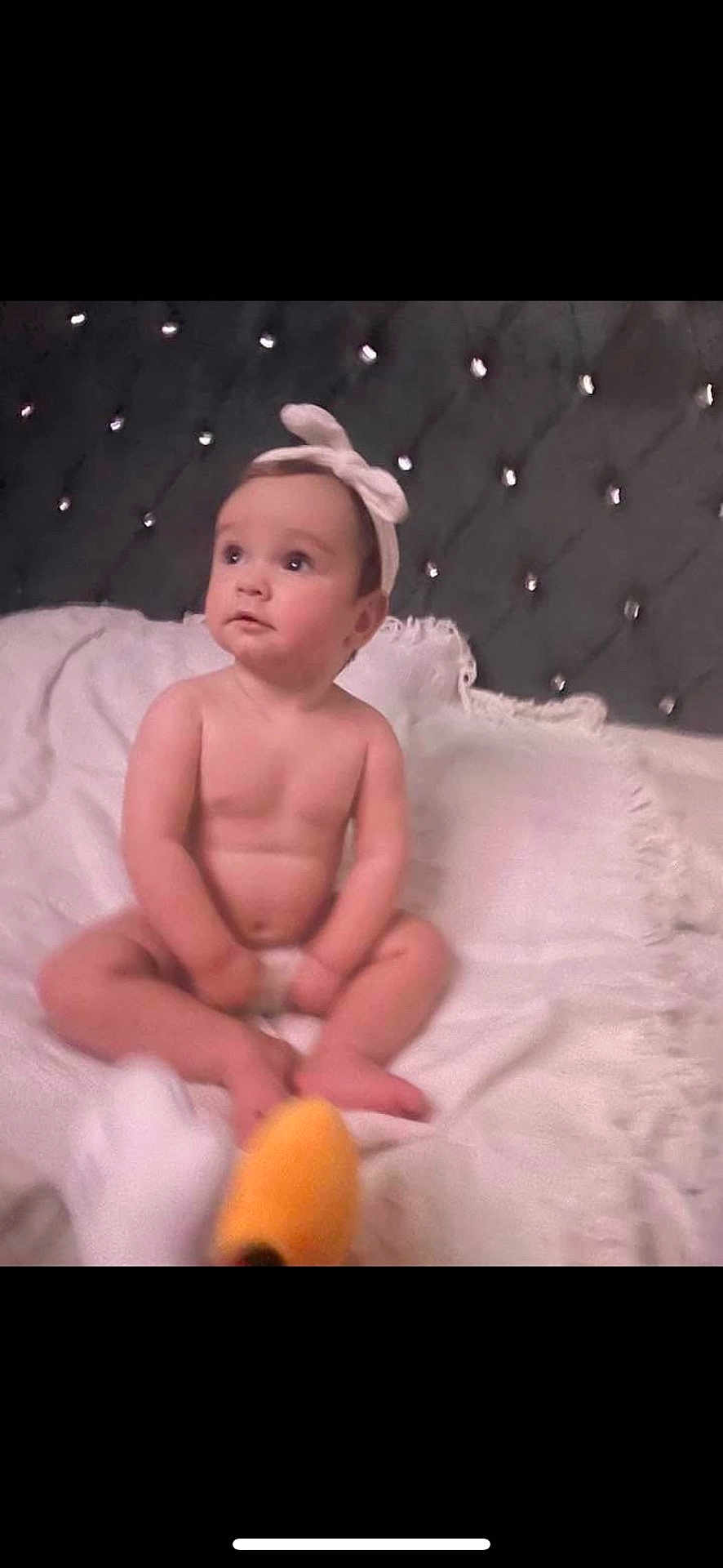 Nevaeh is registered to the contest to win money with this photo: baby, child, headband, blanket, bed, tufted_headboard, plush_toy, soft_texture, indoor, portrait, sitting, curious, infant, white, pink, skin, cute, toy, furniture, cozy