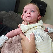 Owen is registered to the contest to win money with this photo: baby, infant, person, hand, clothing, onesie, indoor, cute, holding, cartoon, character, skin, face, expression, newborn, adult_hand, comfort, young, child, portrait