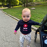Mona is registered to the contest to win money with this photo: toddler, child, park, stroller, sidewalk, grass, trees, sunlight, walking, curious, blue_eyes, flower_pattern, socks, shoes, sweater, nature, outdoor, person, baby, casual_clothing
