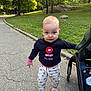 toddler, child, park, stroller, sidewalk, grass, trees, sunlight, walking, curious, blue_eyes, flower_pattern, socks, shoes, sweater, nature, outdoor, person, baby, casual_clothing