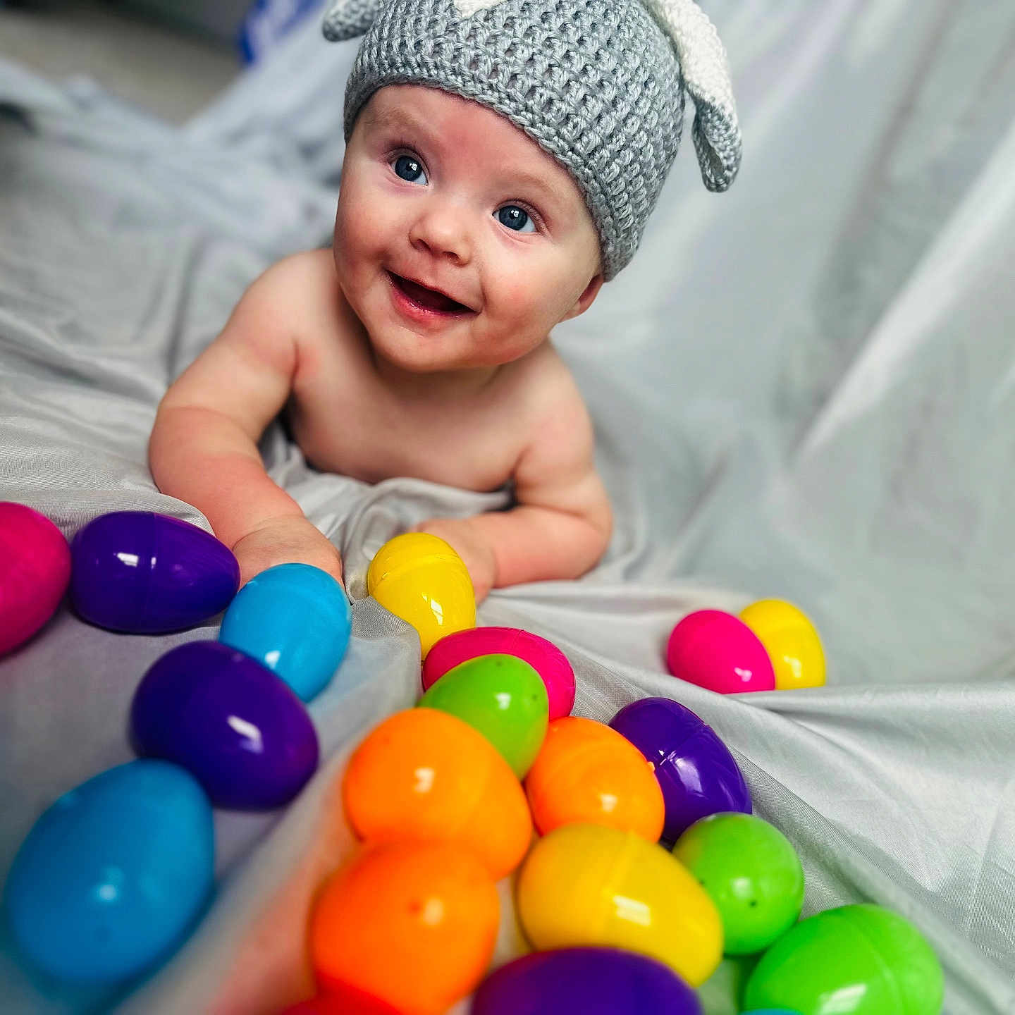 Nova is registered to the contest to win money with this photo: baby, blue_eyes, child, colorful, cute, easter_eggs, face, gray_background, happy, hat, indoors, infant, knitted_hat, lying, plastic_eggs, playful, portrait, sheet, smiling, young_child