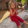 blonde_hair, boulder, child, family_outing, girl, long_hair, outdoor, park, play_area, playground, portrait, red_outfit, rock, shade_sails, sitting, smiling, star_pattern, sunlight, toddler, trees
