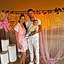 family, baby, man, woman, smiling, indoor, decorations, pink, dress, white_clothing, shoes, floor, flower_garland, lights, curtains, posing, portrait, happy, holding_baby, event