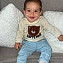 baby, smiling, child, couch, pillow, white_pillow, gray_couch, shirt, bear_design, happy_face_text, pants, socks, indoor, seated, cute, portrait, one_person, infant, happy, cozy