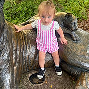 Virginia is registered to the contest to win money with this photo: toddler, child, statue, tiger, bronze, outdoor, greenery, tree, fence, curious, clothing, pink_stripes, shoes, socks, hair, sidewalk, nature, park, person, daylight