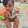baby, child, infant, portrait, smiling, sitting, toy, teething_toy, orange_toy, milestone_sign, last_day_of_school, indoor, classroom, adults, background, blanket, cushion, baby_clothing, dark_hair, family