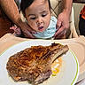 Adrian is registered to the contest to win money with this photo: baby, child, adult_hands, steak, plate, highchair, food, meal, curious, indoor, person, table, dining, appetite, cooking, meat, family, kitchen, expression, closeup