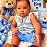 Nestor is registered to the contest to win money with this photo: baby, child, toddler, portrait, sitting, smiling, face, eyes, curly_hair, romper, blue_clothing, shoes, teddy_bear, stuffed_toy, blanket, nursery, bed, indoor, baby_photo, little_boy