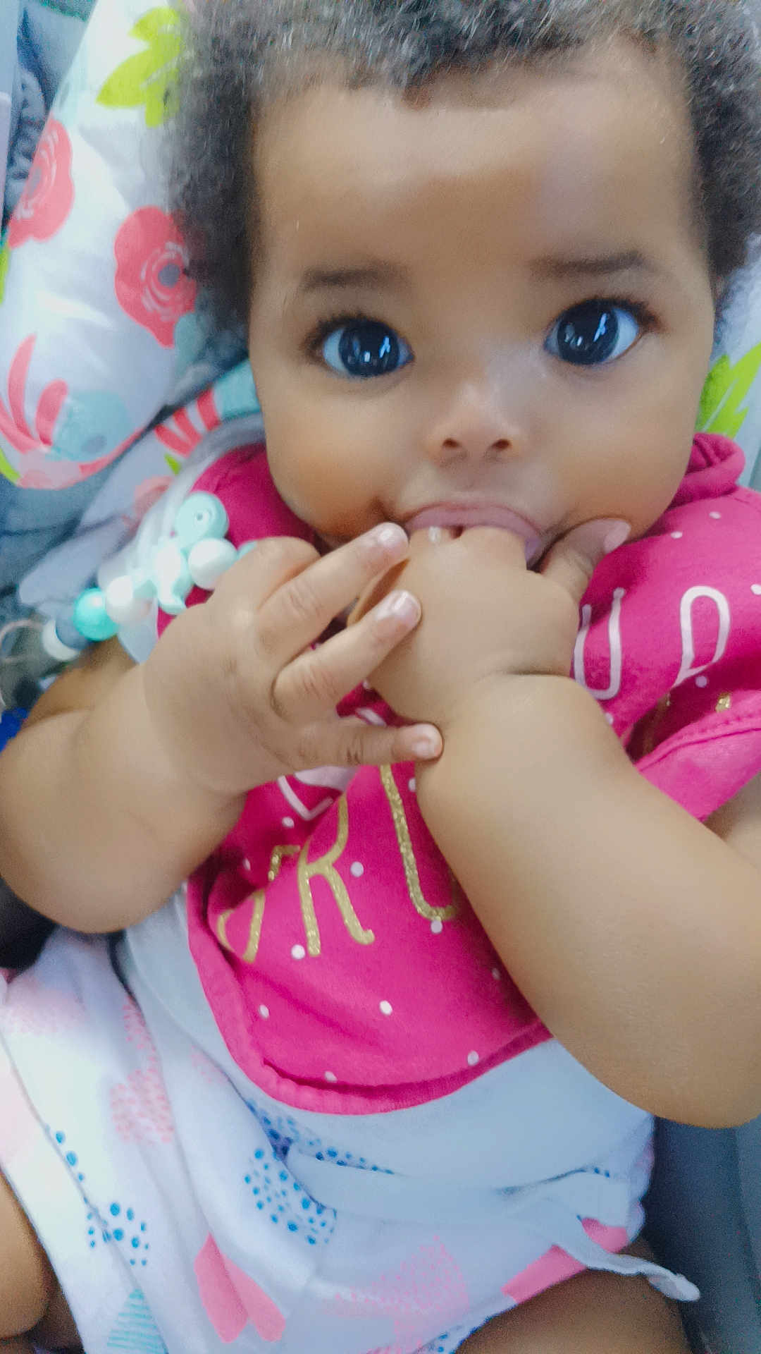 Laylani joined the competition — help win amazing prizes! baby, child, infant, face, hand, bib, clothing, cute, close_up, indoors, patterned_clothes, colorful, skin, person, chewing, sitting, soft_background, floral_pattern, portrait, young