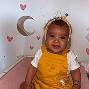 Keliyah a rejoint le concours — aidez-le/la à gagner de superbes lots ! baby, child, smiling, yellow_overall, headband, pink_socks, bracelets, indoor, wall_decor, hearts, stars, moon, cloud, cute, portrait, sitting, infant, happy, face, clothing