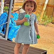 Aphora is registered to the contest to win money with this photo: casual_clothing, child, curly_hair, daytime, denim_dress, expression, fence, green_toy, hat, outdoor, plants, playtime, sippy_cup, sneakers, summer, sunlight, toddler, toy, wooden_deck, young_child