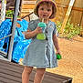toddler, child, hat, denim_dress, sippy_cup, toy, outdoor, wooden_deck, fence, plants, sunlight, curly_hair, sneakers, green_toy, summer, playtime, young_child, casual_clothing, daytime, expression