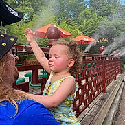 Aphora is registered to the contest to win money with this photo: child, adult, curly_hair, floral_dress, blue_shirt, hat, glasses, outdoor, mister, mist, trees, umbrella, wooden_fence, smile, person, daylight, nature, playful, holding, interaction