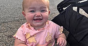 Mclane is registered to the contest to win money with this photo: baby, child, smiling, stroller, outdoor, pink_outfit, pigtails, bows, keychain, happy, cute, infant, person, socks, playful, seated, daylight, building, pavement, expression