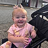 baby, child, smiling, stroller, outdoor, pink_outfit, pigtails, bows, keychain, happy, cute, infant, person, socks, playful, seated, daylight, building, pavement, expression