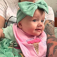 Mclane is registered to the contest to win money with this photo: adult, baby, blue_eyes, bow, closeup, clothing, cute, face, headband, holding, ice_cream_cone, indoor, infant, person, pink_bib, portrait, skin, smile, tattoo, tshirt