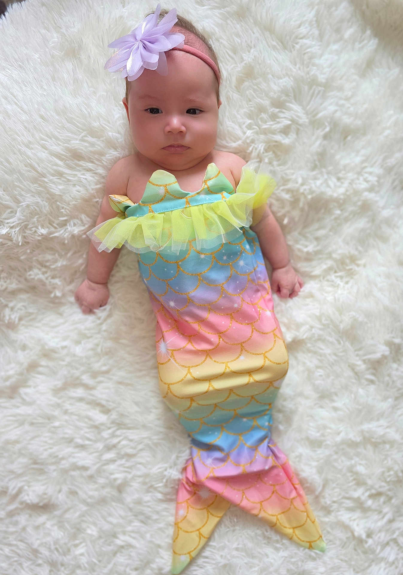 Ella joined the competition — help win amazing prizes! baby, infant, mermaid_costume, headband, flower_headband, pastel_colors, rainbow, lying_down, fluffy_rug, white_rug, cute, child, portrait, costume, soft_texture, indoors, small_child, serious_expression, colorful, adorable