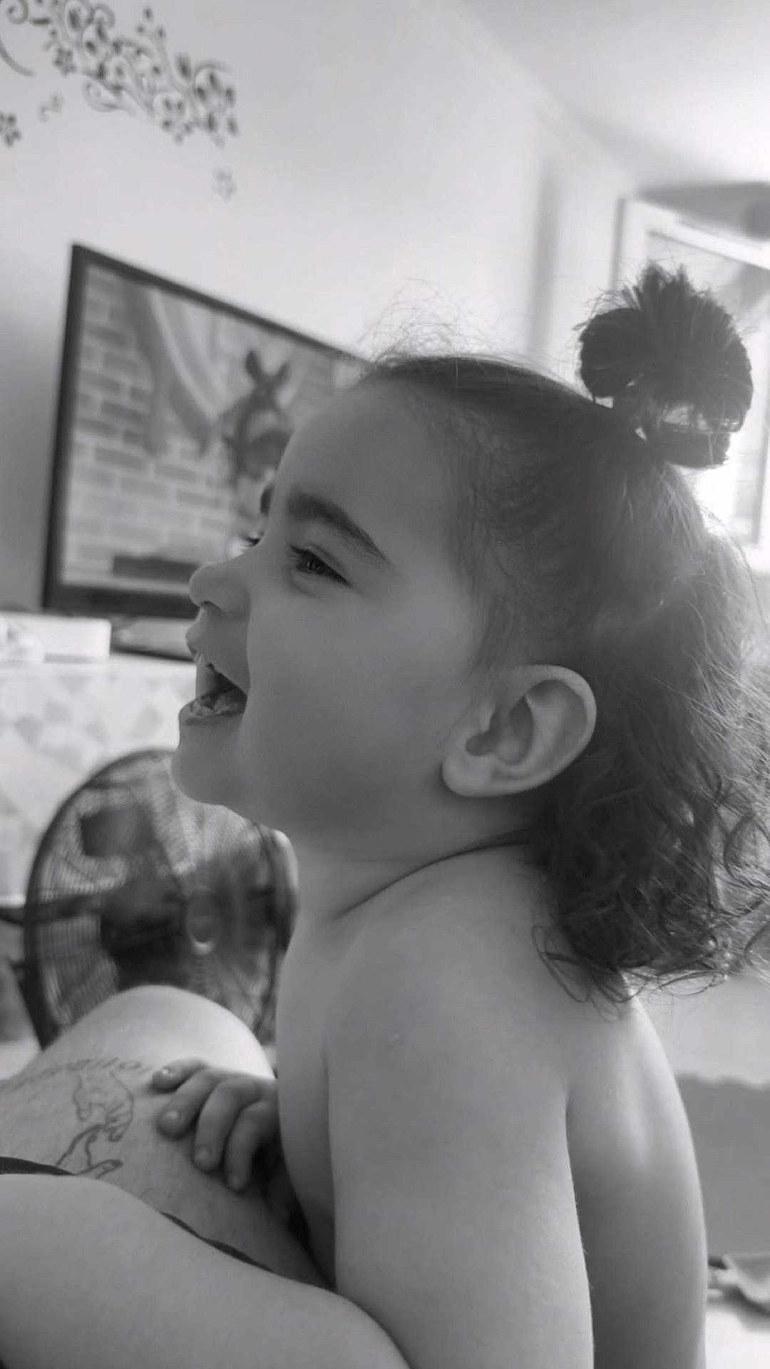 Gioia Maria participe au concours pour gagner de l'argent avec cette photo : arm, black_and_white, bun, child, closeup, curly_hair, fan, furniture, hair, happy, indoor, laughing, person, profile, room, skin, smiling, tattoo, television, toddler