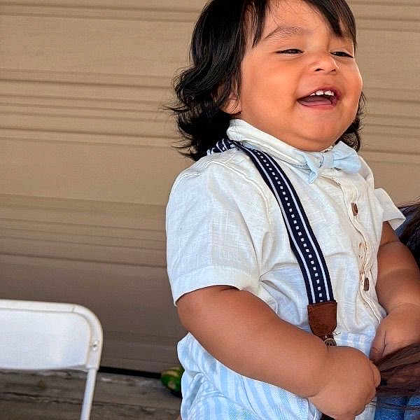 Caleb Paz is registered to the contest to win money with this photo: toddler, child, smiling, happy, white_shirt, suspenders, short_sleeves, dark_hair, indoor, person, cute, young_child, portrait, baby, chubby_cheeks, standing, casual_clothing, background, chair, laughing