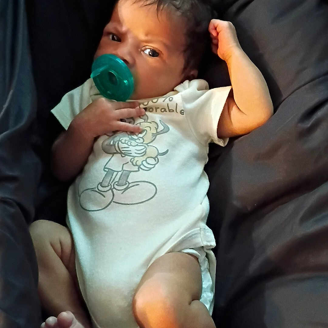 Osiris is registered to the contest to win money with this photo: baby, infant, child, portrait, pacifier, blanket, bed, lying_down, sitting, white_onesie, clothing, dark_background, face, eyes, hair, arm, legs, feet, indoor, relaxed