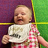 Oakley joined the competition — help win amazing prizes! baby, infant, smile, bib, teddy_bear, pink_clothing, checkered_pattern, carpet, colorful_background, child, cute, happy, lying_down, indoor, young_child, face, person, apparel, text_on_bib, playroom