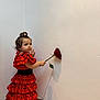 toddler, child, girl, red_dress, polka_dot, flamenco_style, rose, flower, wall, curly_hair, top_knot, indoor, portrait, person, cute, standing, holding, flower_stem, shadow, soft_lighting