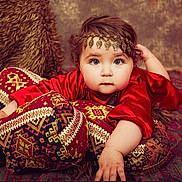 Milena participe au concours pour gagner de l'argent avec cette photo : baby, child, red_clothing, headpiece, cushions, patterned_fabric, carpet, portrait, indoors, decorative, soft_lighting, face, hand, expression, young_child, cozy, texture, traditional, ornamental, furniture