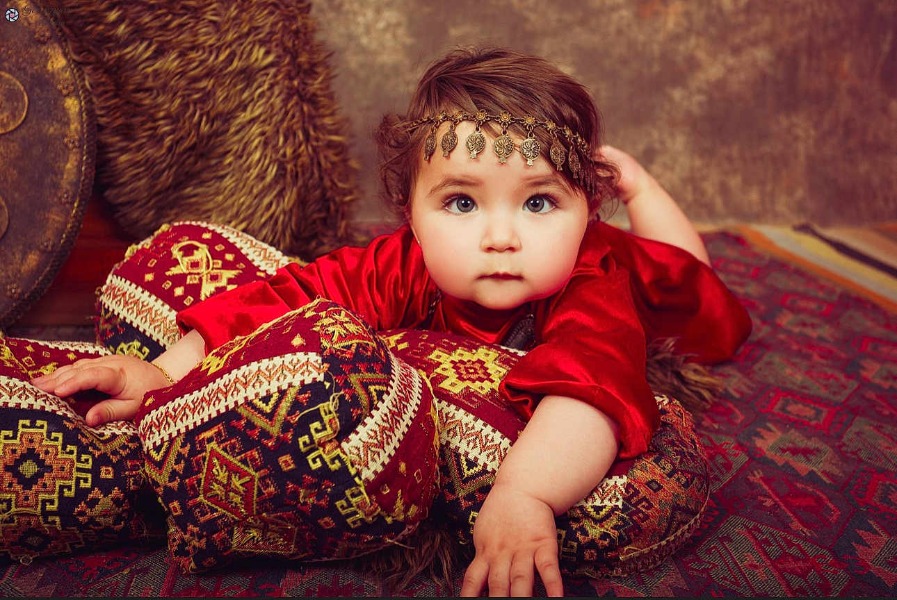 Milena participe au concours pour gagner de l'argent avec cette photo : baby, child, red_clothing, headpiece, cushions, patterned_fabric, carpet, portrait, indoors, decorative, soft_lighting, face, hand, expression, young_child, cozy, texture, traditional, ornamental, furniture