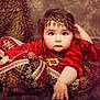 Milena participe au concours pour gagner de l'argent avec cette photo : baby, child, red_clothing, headpiece, cushions, patterned_fabric, carpet, portrait, indoors, decorative, soft_lighting, face, hand, expression, young_child, cozy, texture, traditional, ornamental, furniture