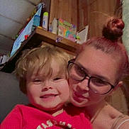 Levi is registered to the contest to win money with this photo: child, woman, glasses, smile, red_tshirt, dinosaur_graphic, indoor, wood_paneling, shelf, toiletries, hair_bun, casual_clothing, portrait, happy, family, closeup, person, holding, background, home