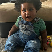 Tripp joined the competition — help win amazing prizes! baby, child, toddler, curly_hair, portrait, sitting, indoors, living_room, couch, carpet, denim_overalls, green_shirt, toy, colorful_toy, floor, home, looking_at_camera, facial_expression, natural_light, photography