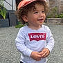 building, cap, casual_clothing, child, curly_hair, curly_hair_kid, daylight, fence, gravel, happy, levis, orange_cap, outdoor, plants, portrait, smile, standing, toddler, white_shirt, young_child