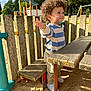 Eymen a rejoint le concours — aidez-le/la à gagner de superbes lots ! bench, casual_clothing, child, curly_hair, daytime, fence, happy, nature, outdoor, park, person, playground, sand, smiling, sneakers, striped_shirt, sunlight, toddler, trees, wooden_table