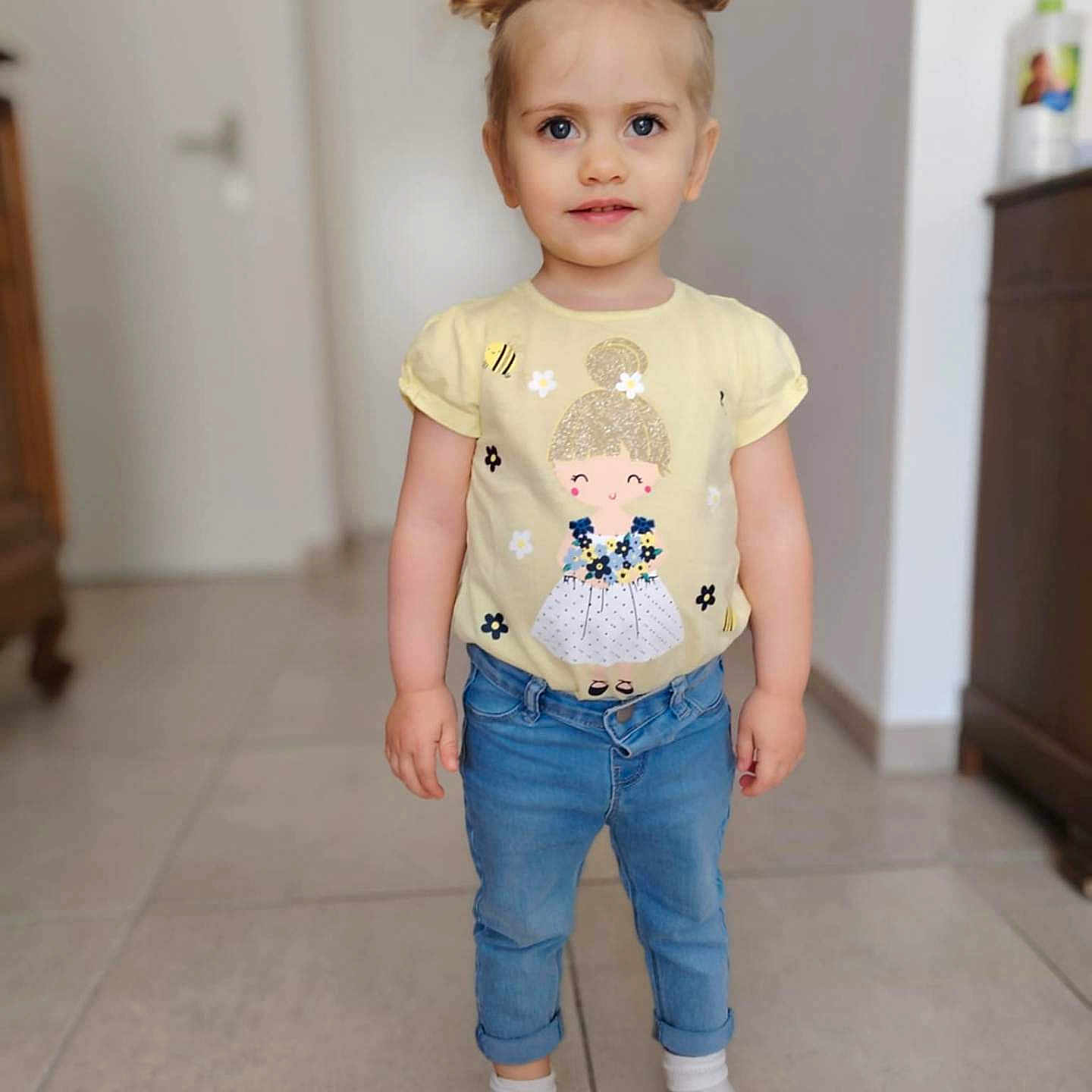 Torvi participe au concours pour gagner de l'argent avec cette photo : casual_wear, child, clothing, curious, cute, face, floor, hair_buns, home_interior, indoor, jeans, person, portrait, smile, socks, standing, tile_floor, toddler, yellow_shirt, young_child