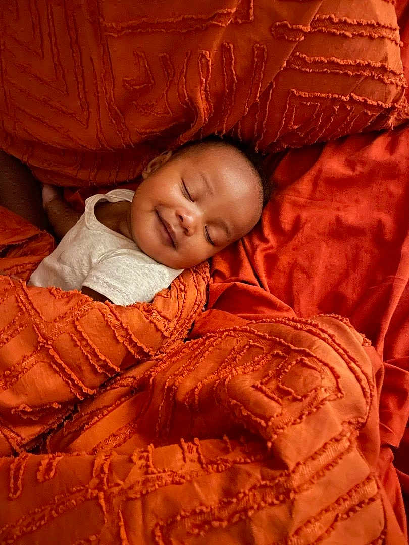 Jalen joined the competition — help win amazing prizes! baby, sleeping, smile, blanket, orange, texture, bed, comfort, rest, peaceful, infant, face, person, young_child, cozy, soft, relaxation, indoor, cushion, cute