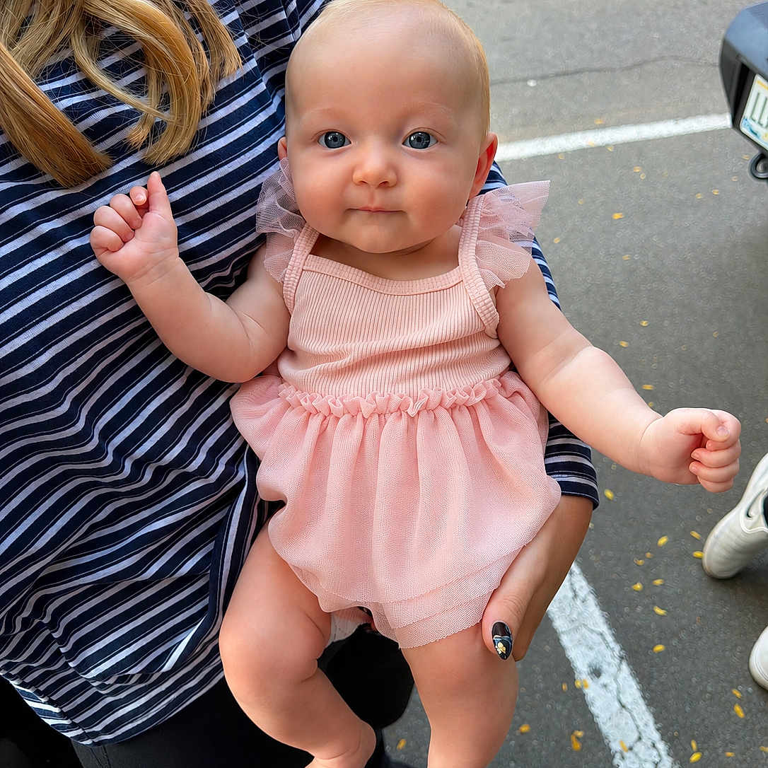Adelyn joined the competition — help win amazing prizes! baby, child, person, holding, pink_dress, striped_shirt, outdoor, parking_lot, blue_eyes, curly_hair, cute, infant, hand, foot, skin, smile, casual_clothing, daylight, human, portrait