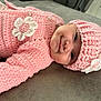 baby, infant, pink_clothing, crocheted_hat, crocheted_sweater, flower_decoration, smiling, face, headwear, lying_down, soft_surface, close_up, cute, child, portrait, indoor, warm_clothing, happy, small_person, adorable