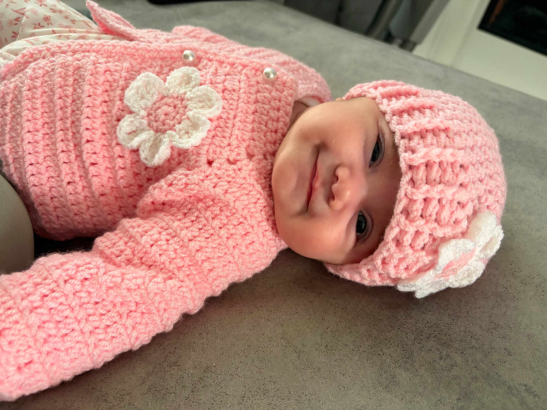 Alyzia-Mae is registered to the contest to win money with this photo: baby, infant, pink_clothing, crocheted_hat, crocheted_sweater, flower_decoration, smiling, face, headwear, lying_down, soft_surface, close_up, cute, child, portrait, indoor, warm_clothing, happy, small_person, adorable