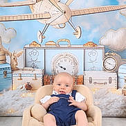 Gaspard participe au concours pour gagner de l'argent avec cette photo : baby, child, infant, armchair, overalls, blue_clothing, smiling, face, indoor, wood_floor, background, decor, toy_plane, clock, clouds, cute, portrait, seated, happy, studio