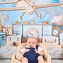 baby, child, infant, armchair, overalls, blue_clothing, smiling, face, indoor, wood_floor, background, decor, toy_plane, clock, clouds, cute, portrait, seated, happy, studio