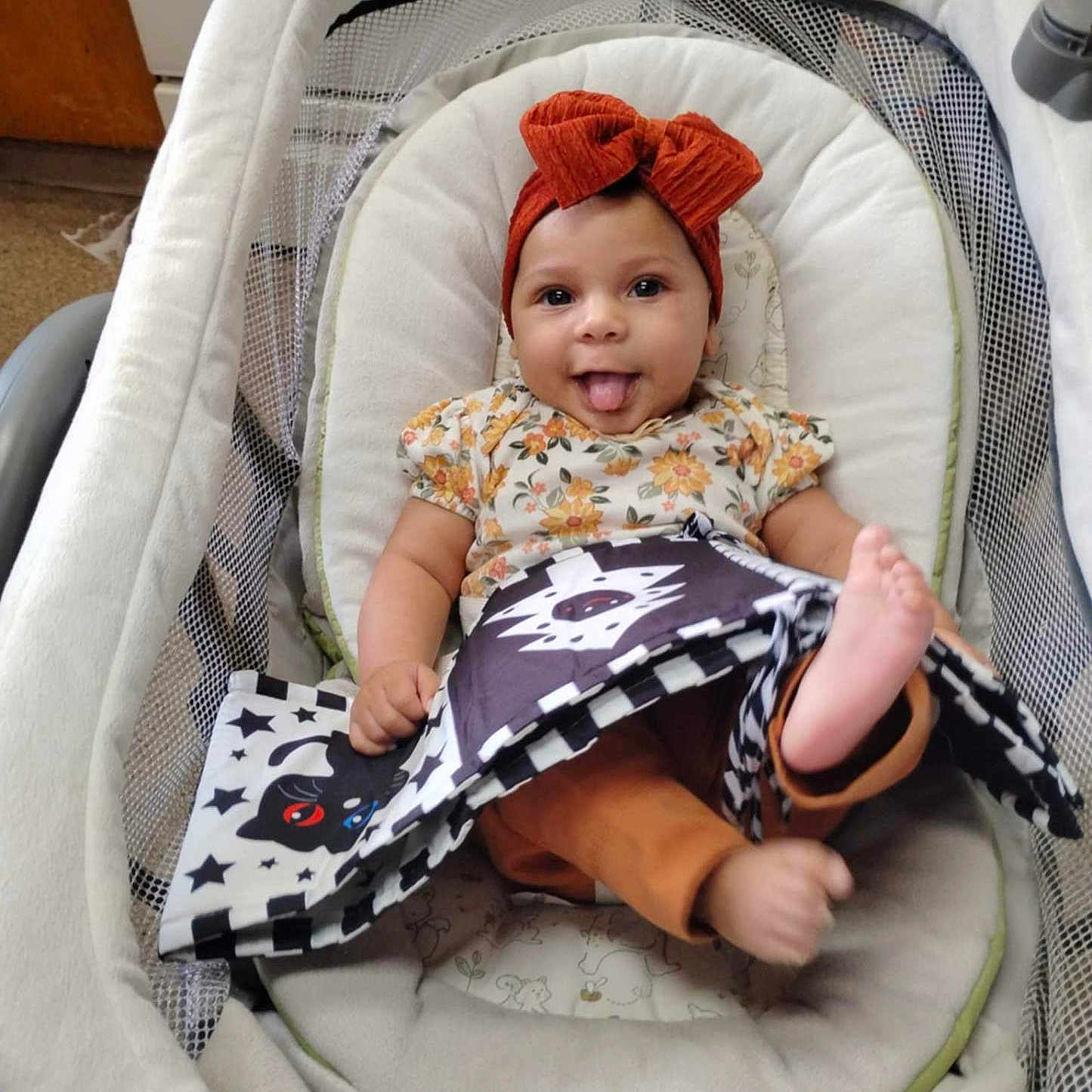 Gloria joined the competition — help win amazing prizes! animal_pattern, baby, baby_seat, black_and_white_book, bow, carpet, child, cute, floral_clothing, happy, headband, indoors, infant, orange_pants, person, playful, portrait, smiling, soft_cushion, tongue_out