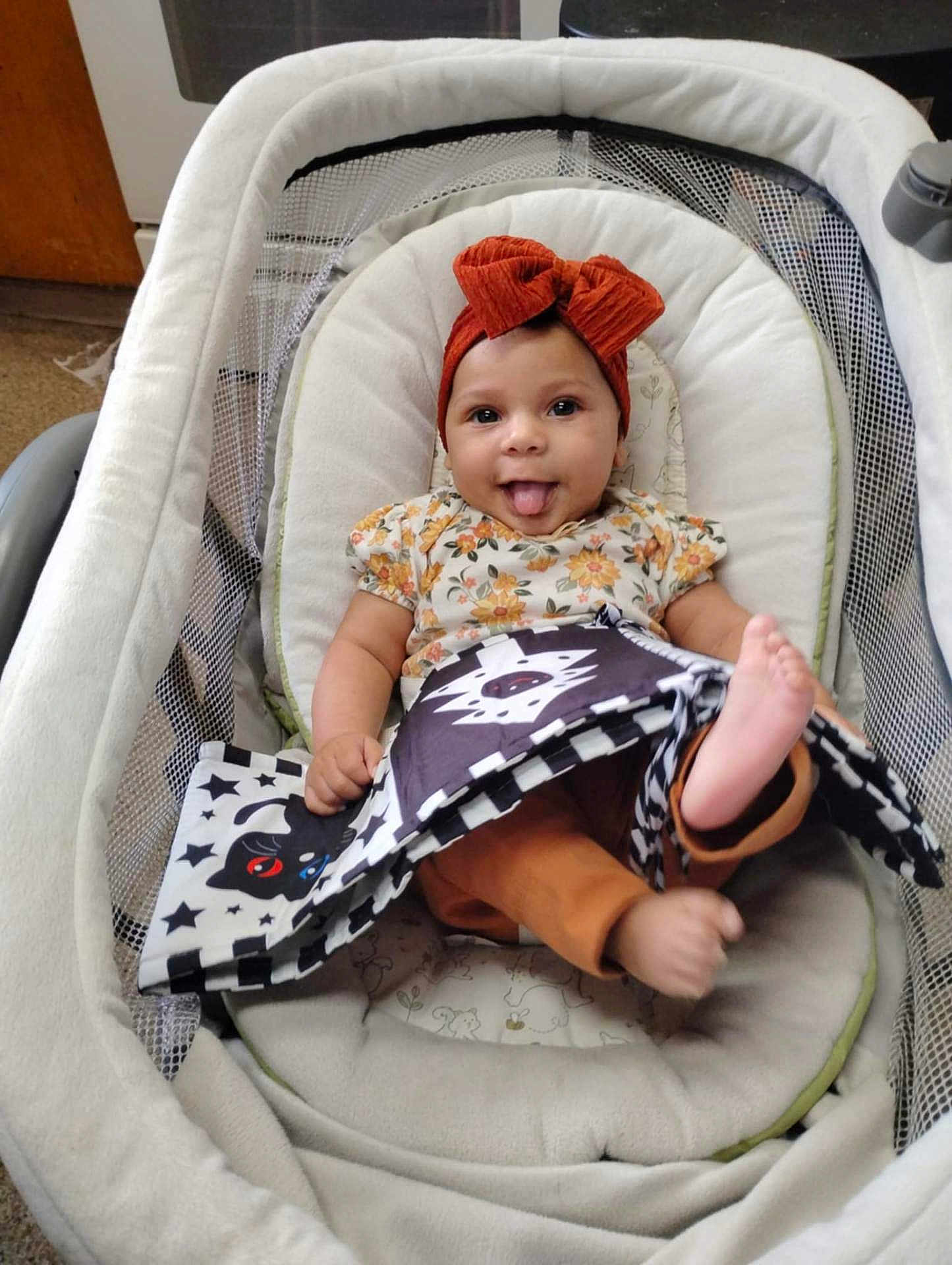Gloria joined the competition — help win amazing prizes! baby, infant, child, headband, bow, tongue_out, baby_seat, soft_cushion, floral_clothing, orange_pants, black_and_white_book, animal_pattern, indoors, carpet, happy, playful, cute, smiling, person, portrait