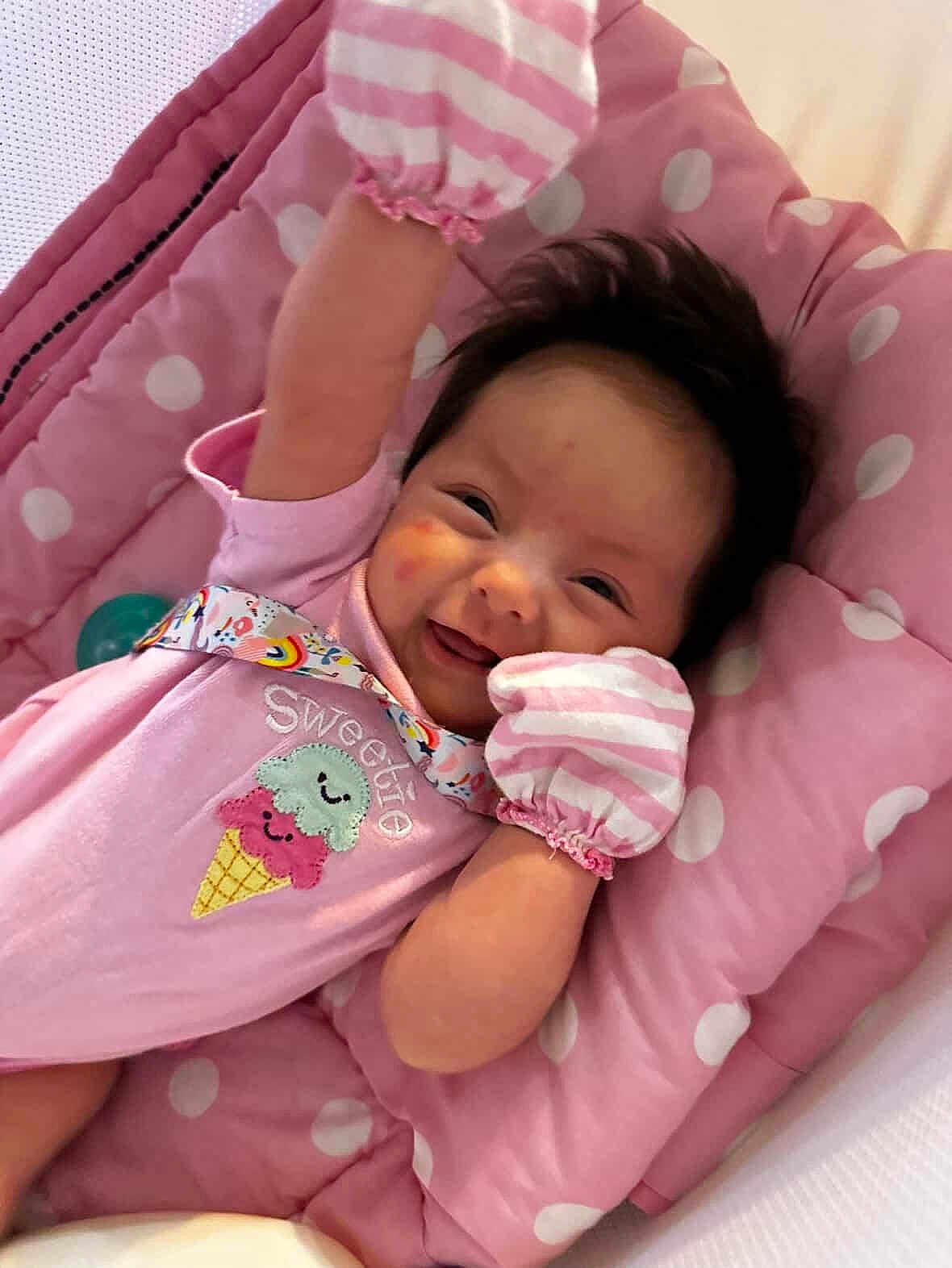 Madison is registered to the contest to win money with this photo: baby, smiling, pink, mittens, polka_dot, cushion, infant, cute, happy, lying_down, clothing, face, portrait, soft_toys, indoors, child, sweetie, ice_cream, striped, handwear