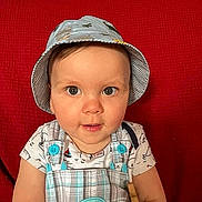 Raphaël participe au concours pour gagner de l'argent avec cette photo : baby, bucket_hat, car_patch, child, clothing, curious, cute, eyes, face, hat, indoor, infant, overalls, person, plaid, portrait, red_background, sitting, smiling, young_child