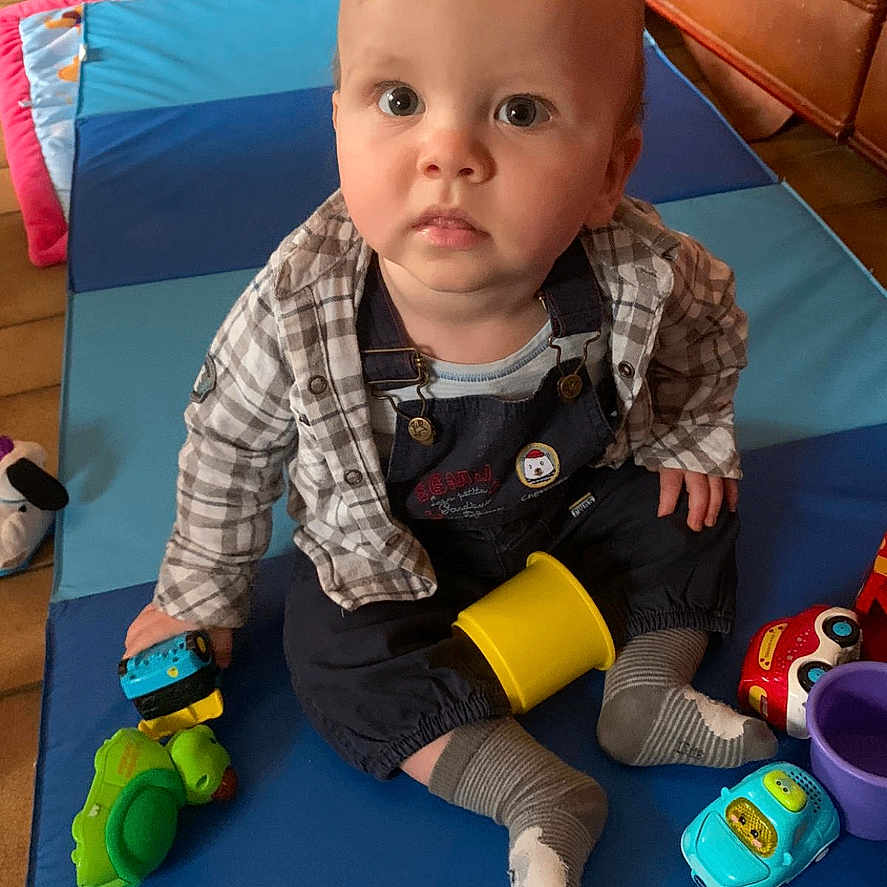 Raphaël a rejoint le concours — aidez-le/la à gagner de superbes lots ! baby, casual_clothing, child, colorful, couch, cup, curious, expression, floor, indoor, looking_up, mat, overalls, plastic_toys, playing, seated, shirt, socks, toddler, toy